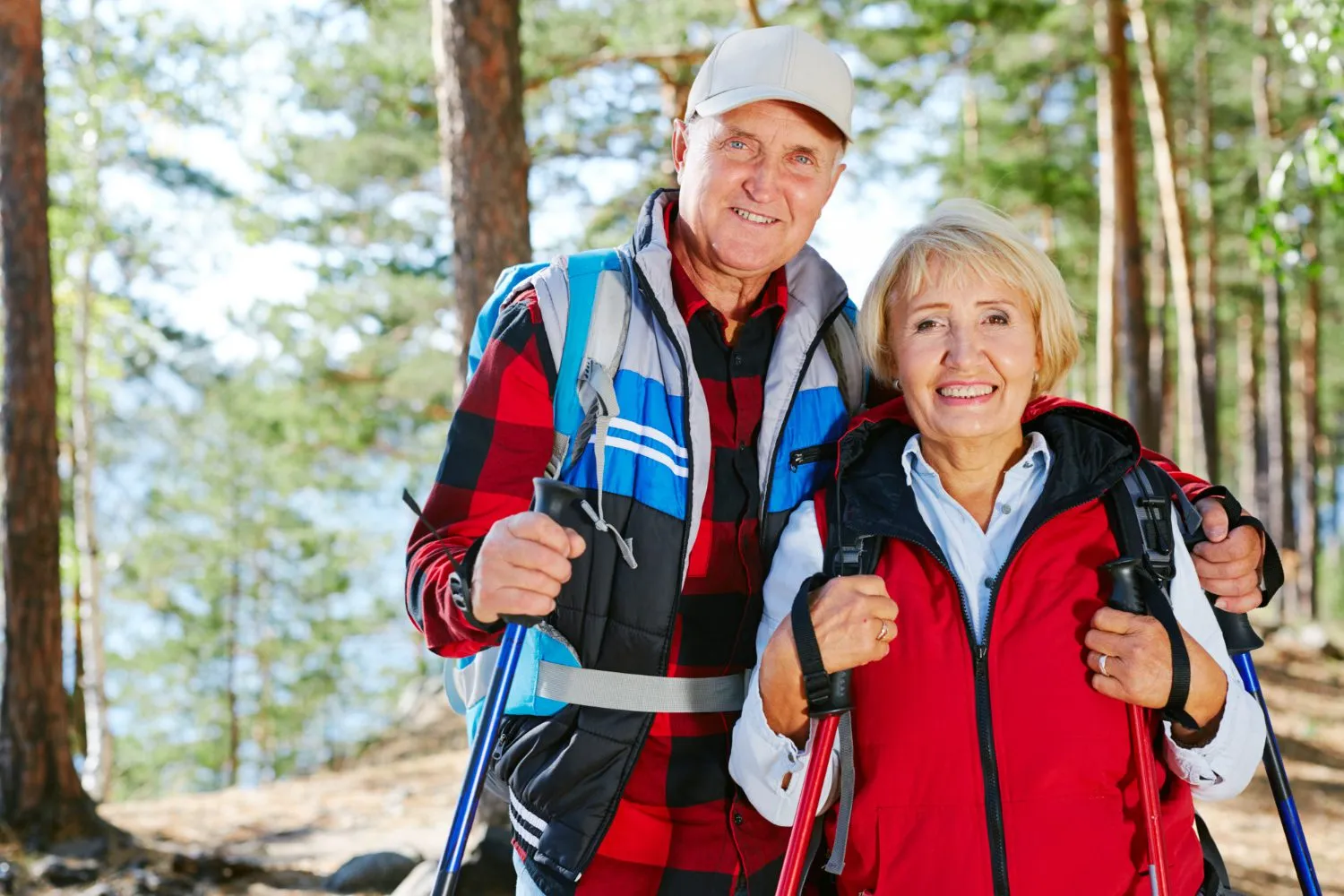 Two hikers in colorful jackets stand together in a sunlit forest, holding trekking poles, surrounded by tall trees
